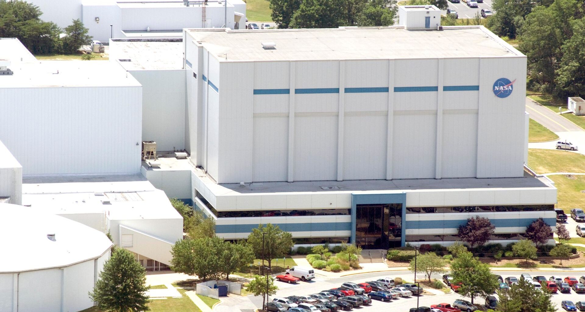 A series of white buildings are seen on a hillside with a parking lot full of cars in the front. One of the buildings has the blue NASA meatball logo on it