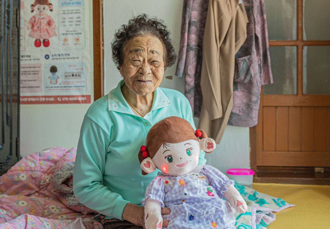 Elderly woman holds an AI-powered Hyodol plushie robot companion. The device is designed to spend 24 hours a day with people who live alone, providing emotional support and health care monitoring.