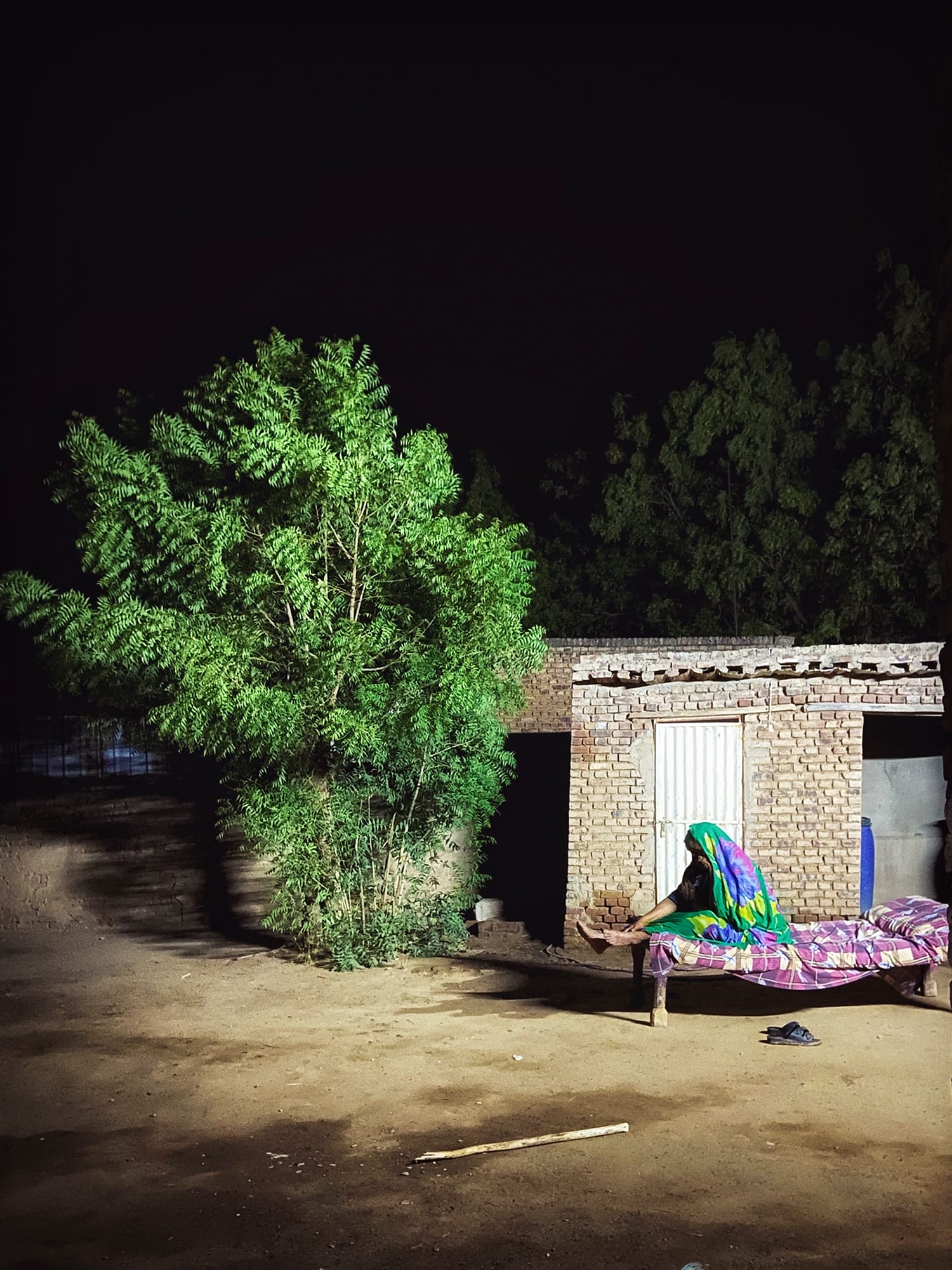 a photograph by Jood Elshiek of a woman sitting on a cot outside of a small stone home, lit up at night