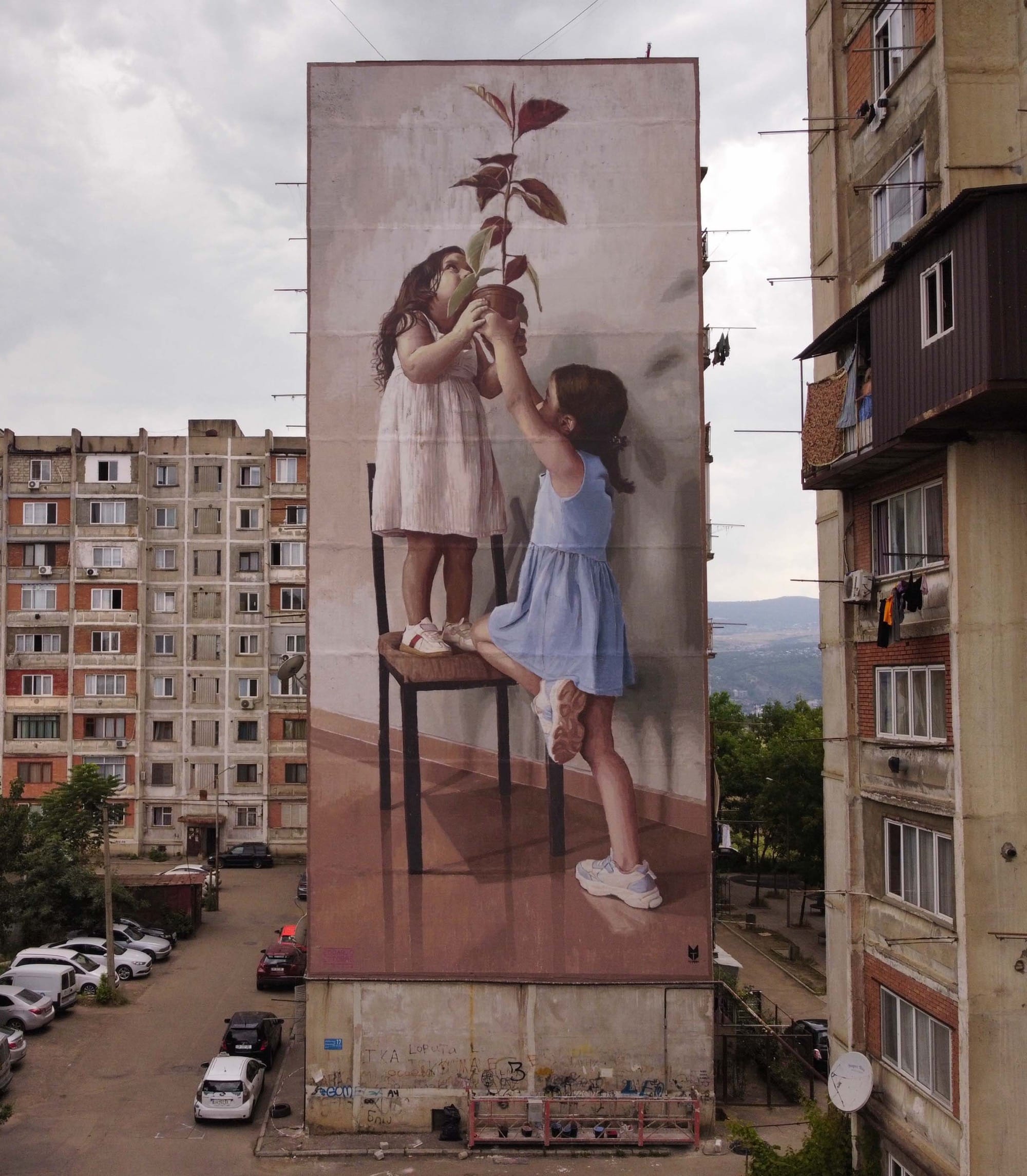 a mural on the side of a tall building showing two young girls playing around on a chair, holding a potted plant