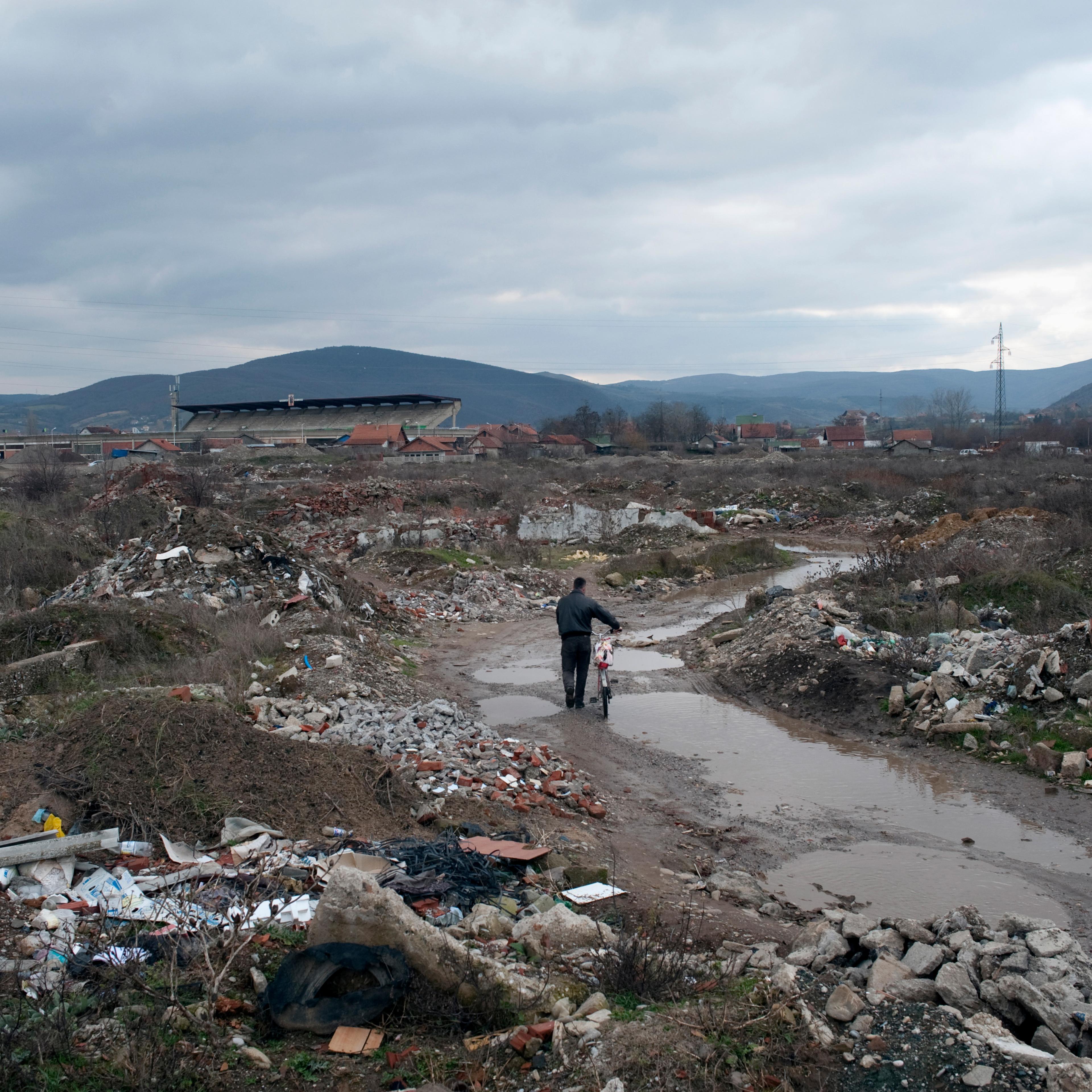 A person pushing a bike through a muddy landfill with debris under an overcast sky, distant hills and houses.
