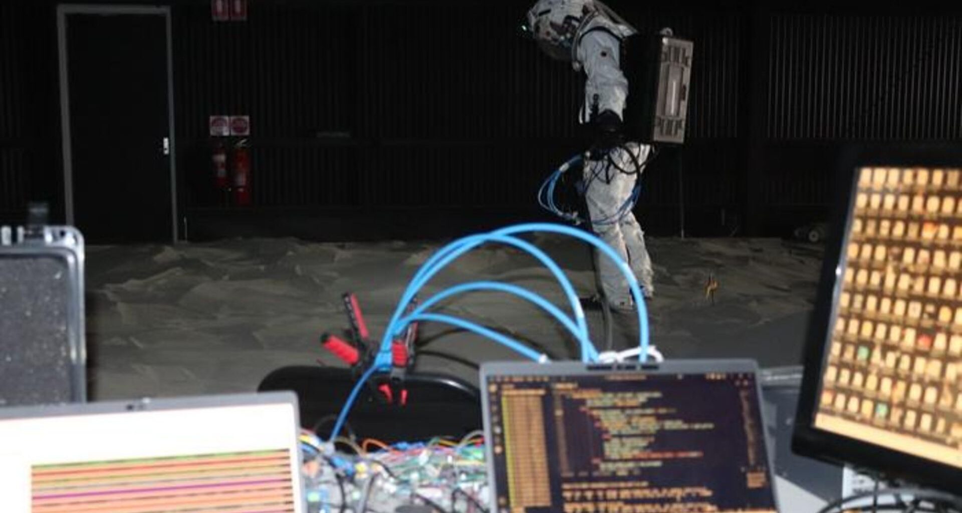 A person wearing a white suit stands behind a desk with various wires and computers on it