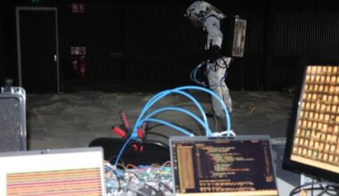 A person wearing a white suit stands behind a desk with various wires and computers on it