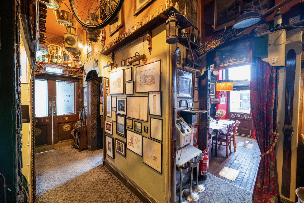 Interior of The Canny Man's pub, a narrow hall with framed pictures, historical items, and doors leading to a dining area and possibly "The Volunteer Arms."