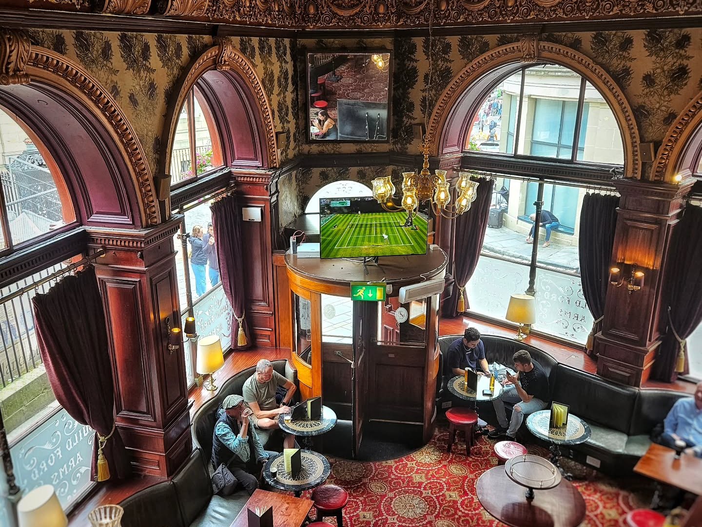 A pub with ornate wooden arches, a red patterned carpet, and people sitting at tables.