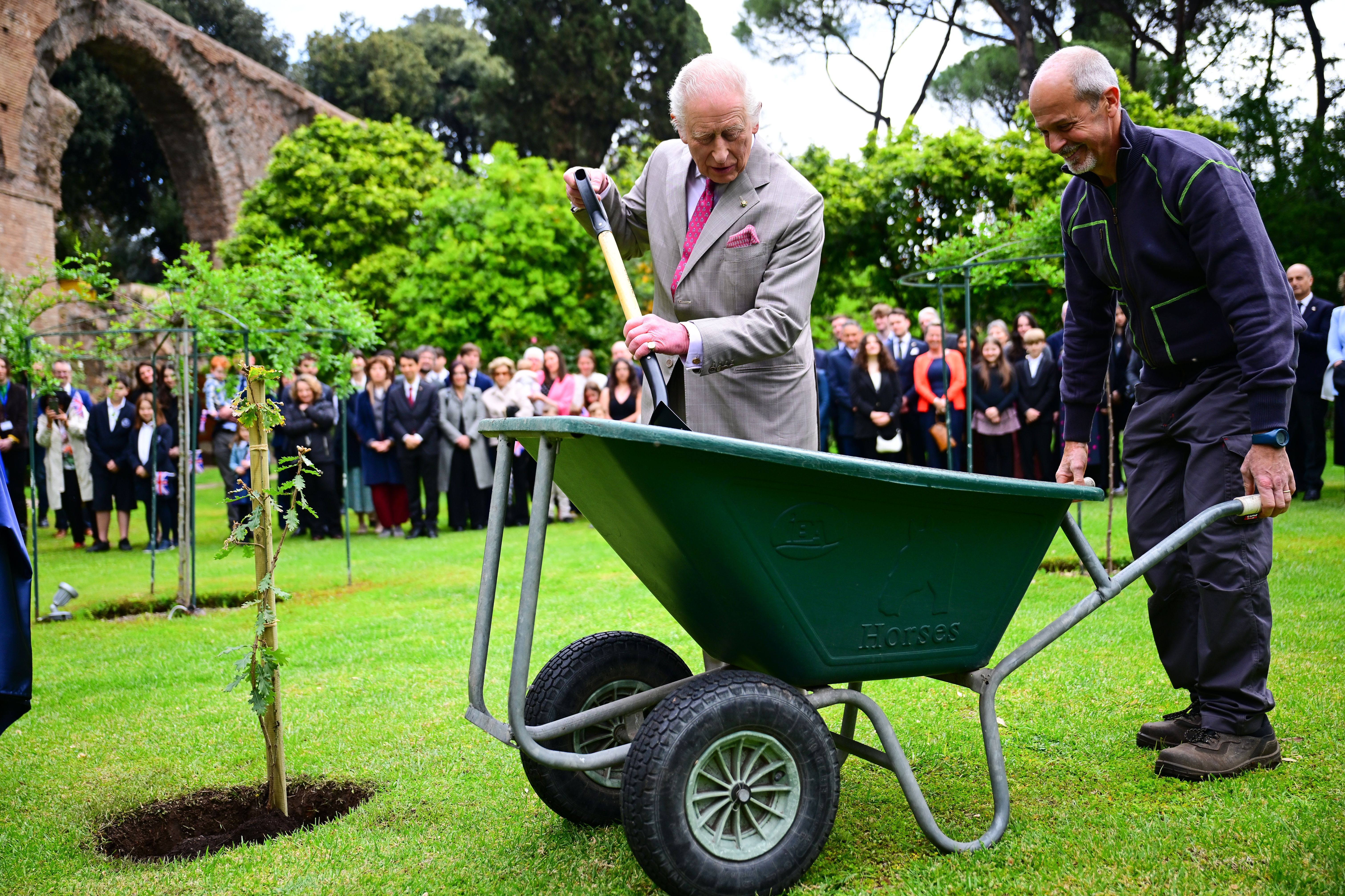 King Charles III planting a tree with a shovel while a man holds a wheelbarrow.