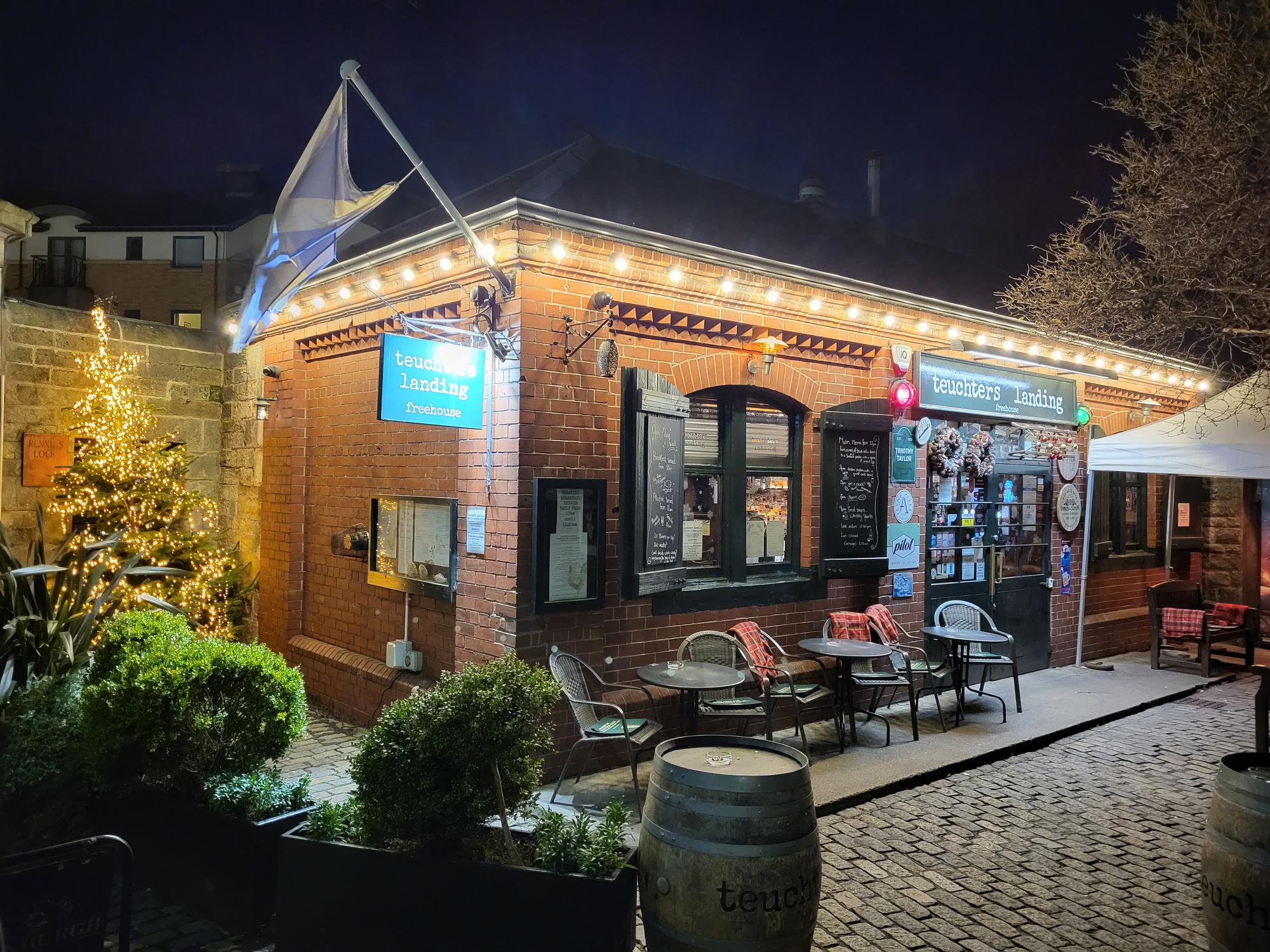 Teuchters Landing pub at night, with Christmas decorations including a lit Christmas tree and lights strung along the building.