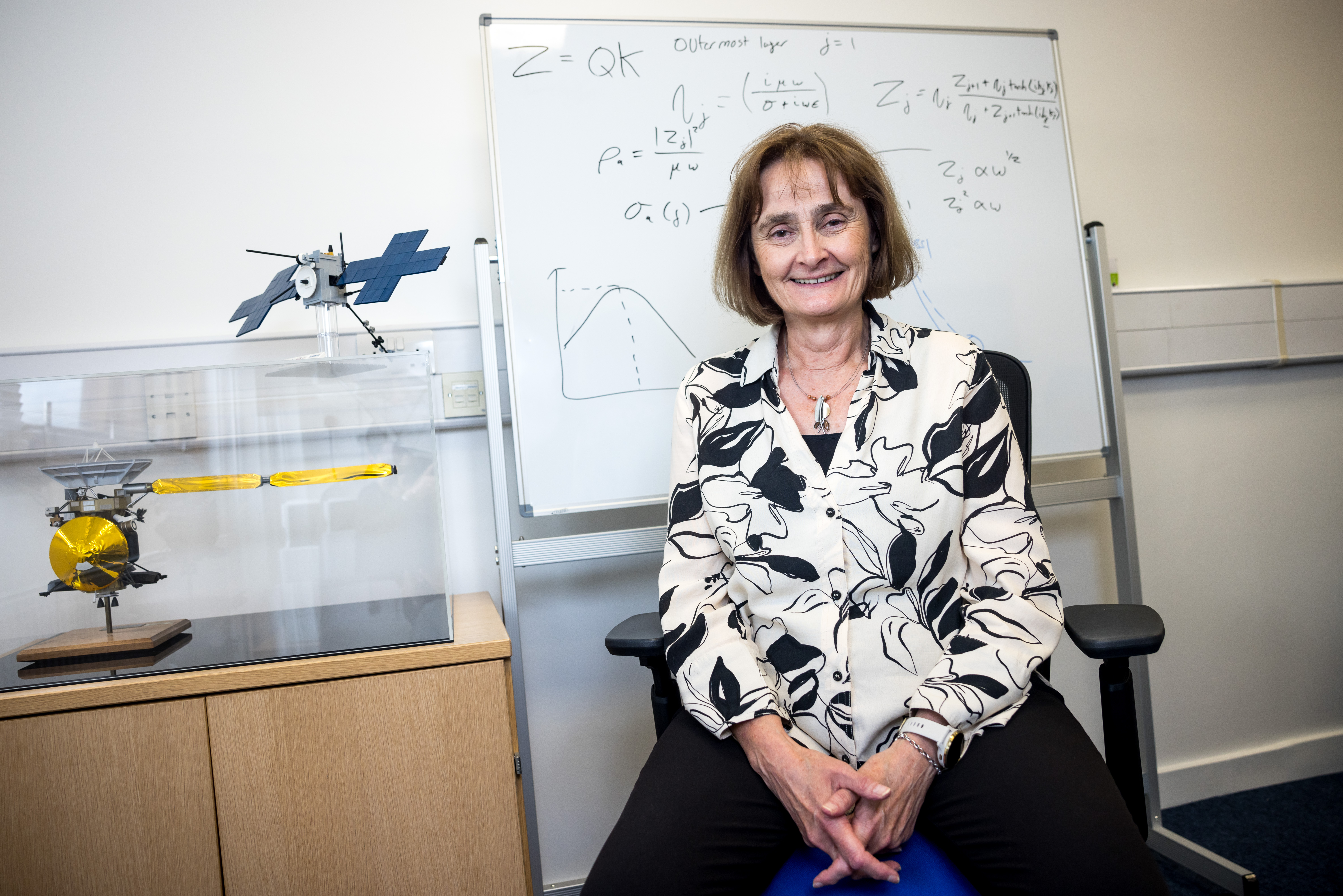 Professor Michele Dougherty, Astronomer Royal, sits in her university office with a whiteboard of physics equations and models of spacecraft.