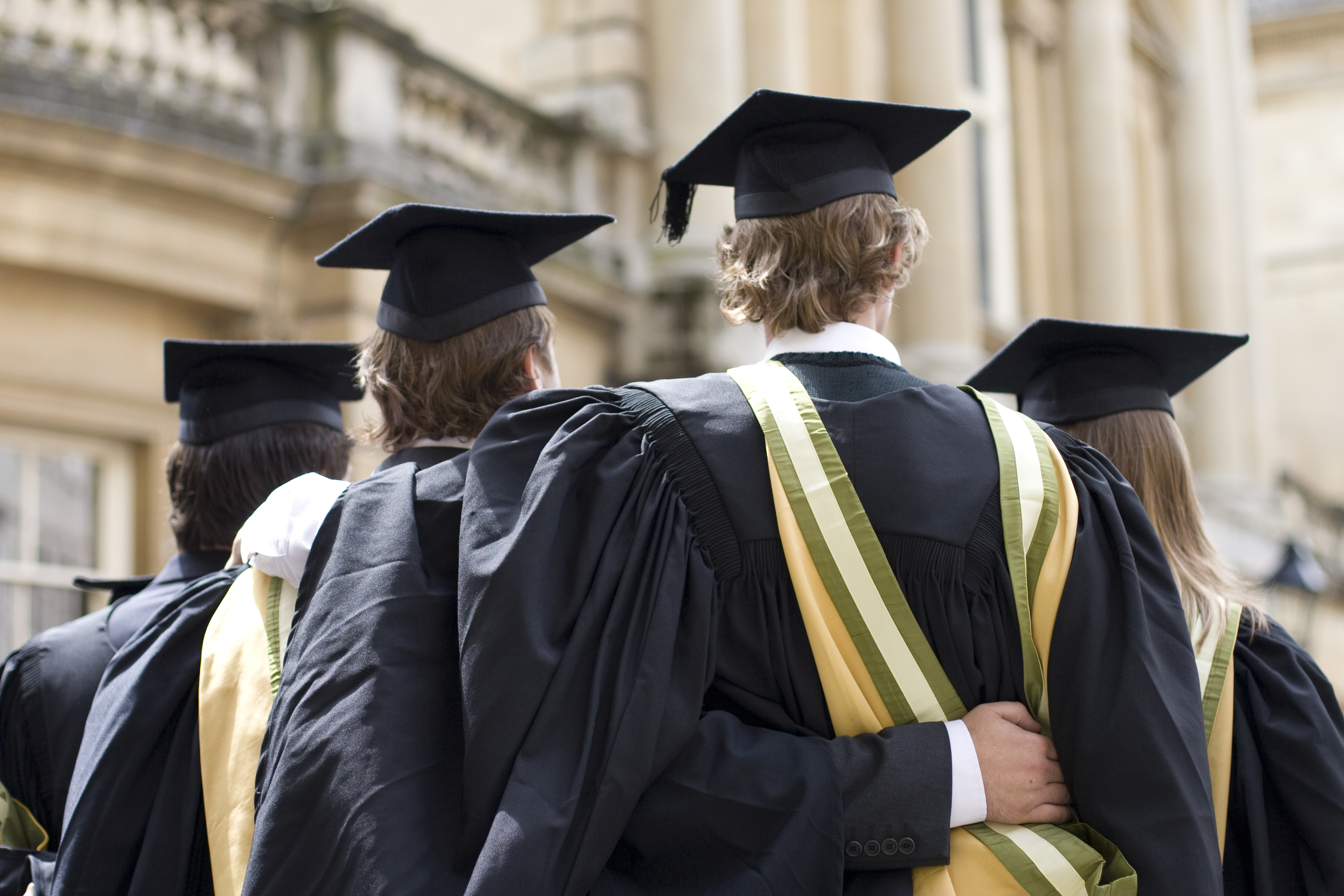 Four university students in graduation gowns and mortarboards, seen from behind, standing side-by-side, with one student's arm around another.