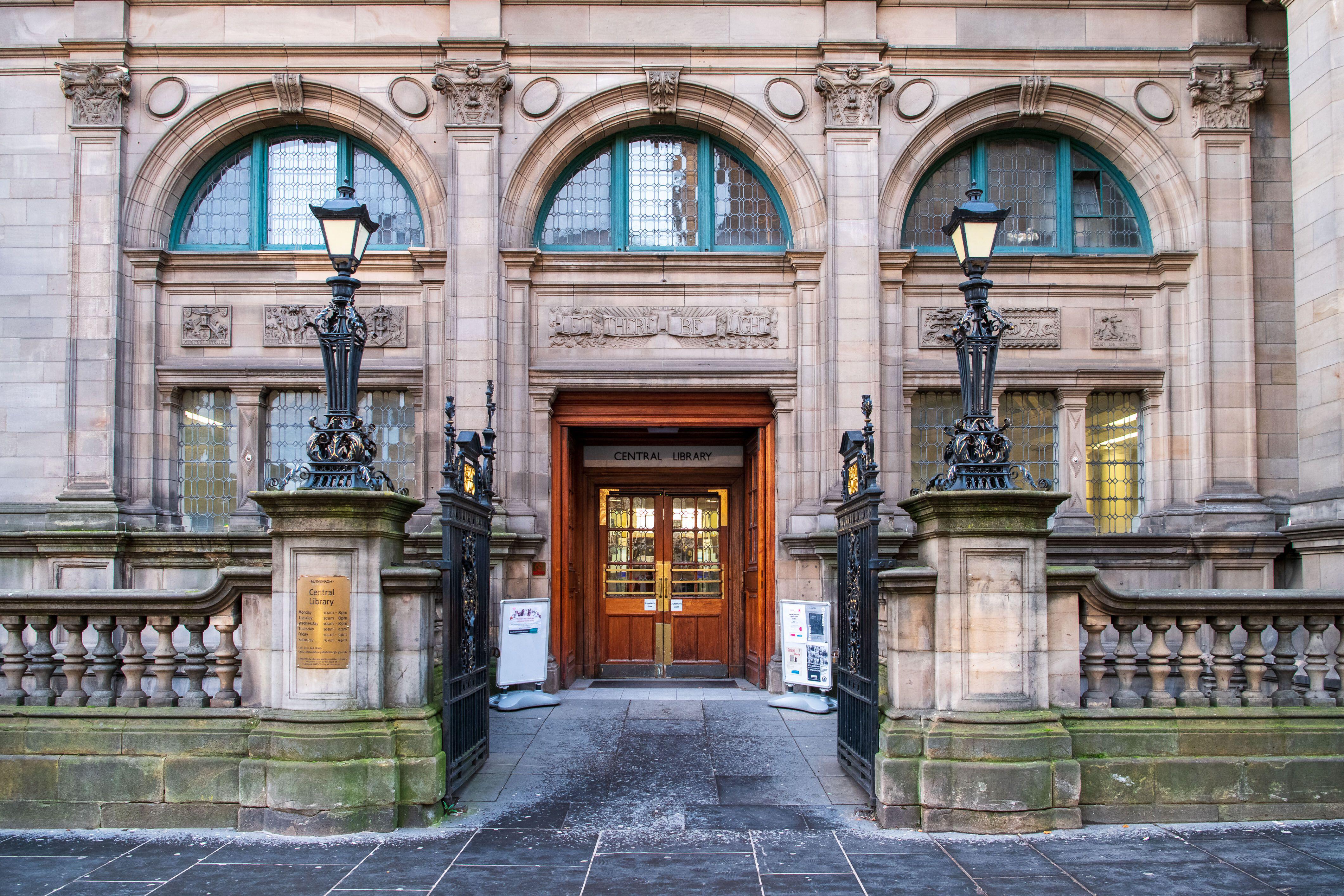 The entrance to the Central Library with ornate stone carvings, large arched windows, two tall street lamps, and a wooden double door with a "Central Library" sign above it.