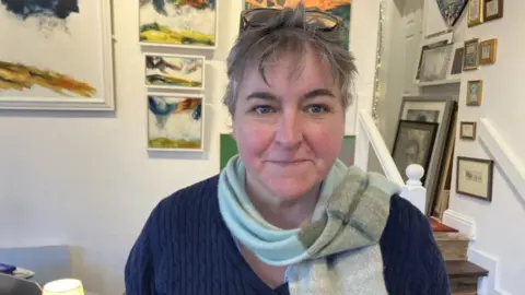A woman has glasses perched on her head, and a scarf around her neck. Behind her there are a number of paintings on the wall