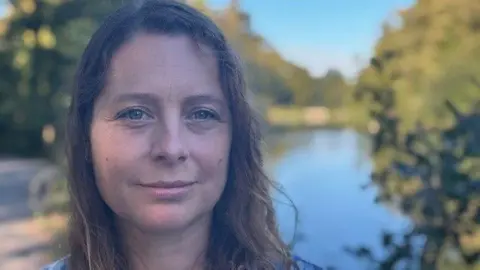 Hannah Cloke, with wavy, dark hair, can be seen looking straight into the camera. Behind her is a large pond and nature area at the University of Reading.