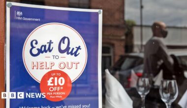 An Eat Out to Help Out poster in a window with two wine glasses and a blurred reflection of a customer