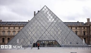 The outside of the glass pyramid at the Louvre Museum in Paris on a grey day, with personnel outside, one of whom is wearing a high-vis jacket