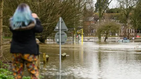 Getty Images A woman can be seen from behind holding a phone and taking pictures of submerged cars in a car park after heavy rains and sewer system overflows caused the River Thames to break its banks, on 5 January 2024. She is wearing a blue fleece and floral trousers - she also has died blue hair.
