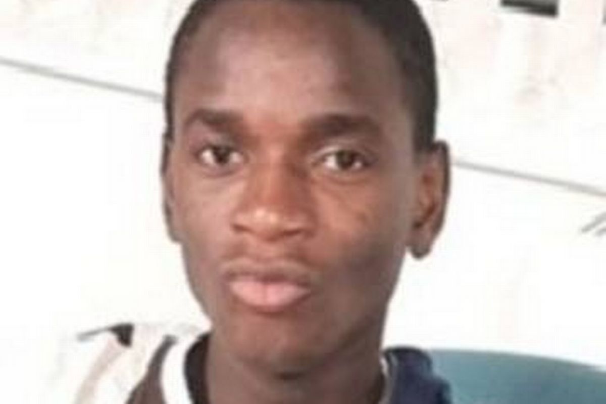 An individual with short hair and a serious expression, seated against a plain background, possibly in an indoor setting.