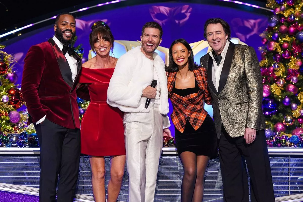 Five individuals dressed in formal attire are standing together on a stage, possibly for a televised event. One person in the center holds a microphone, while others stand beside them, facing the camera. The background features vibrant colors and decorative elements.