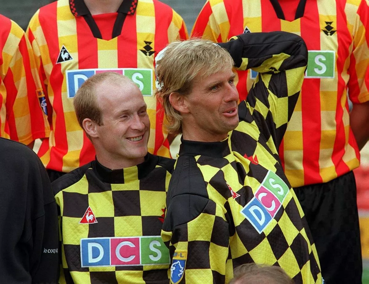 Two individuals in yellow and black checkered jerseys, standing together with several other similarly dressed individuals in the background, possibly at a sporting event.