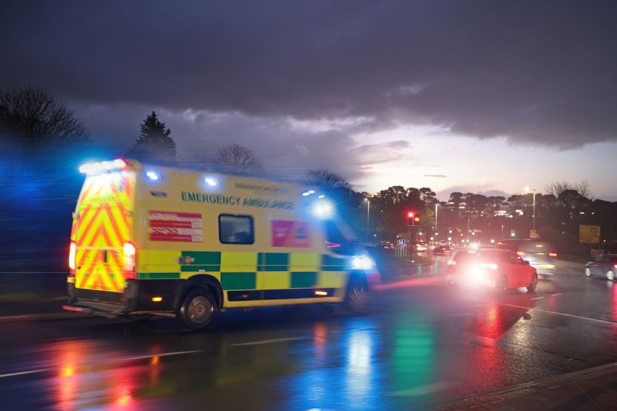 Ambulance driving on road at dusk on wet road with emergency lights and siren on.