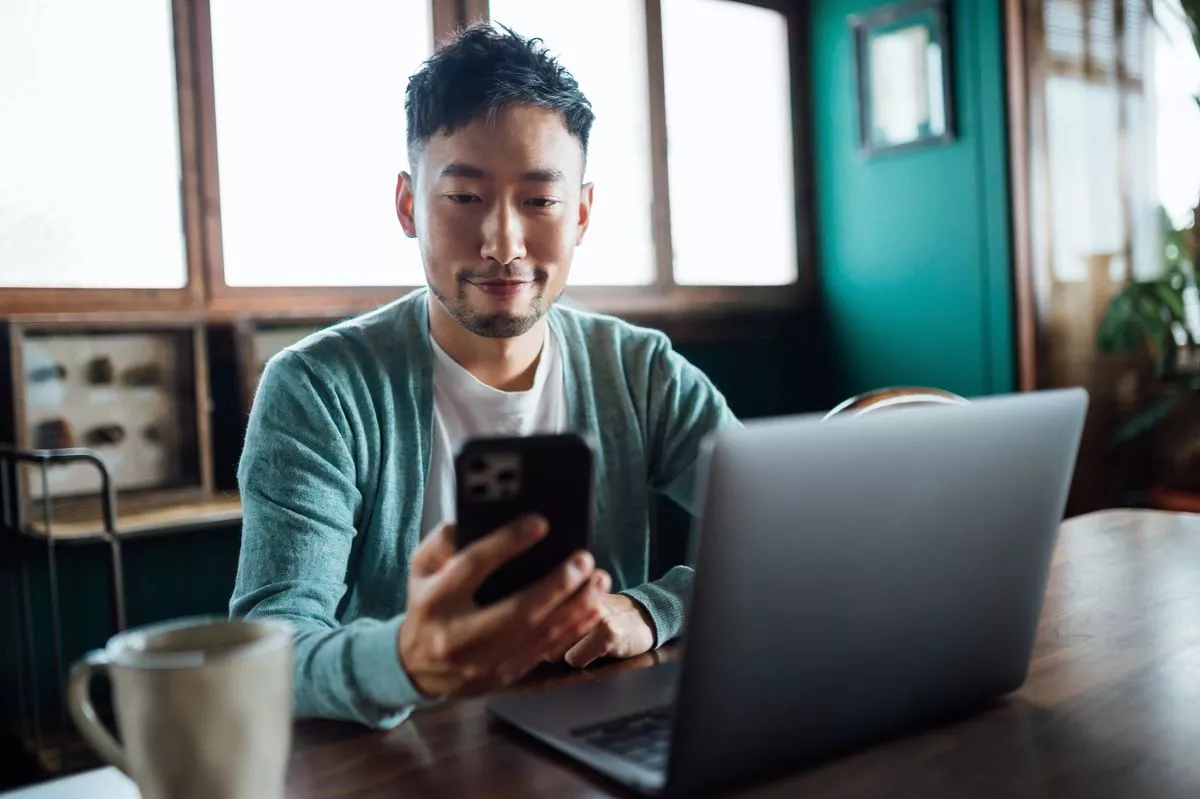 Confident young Asian man looking at smartphone while working on laptop computer in home office
