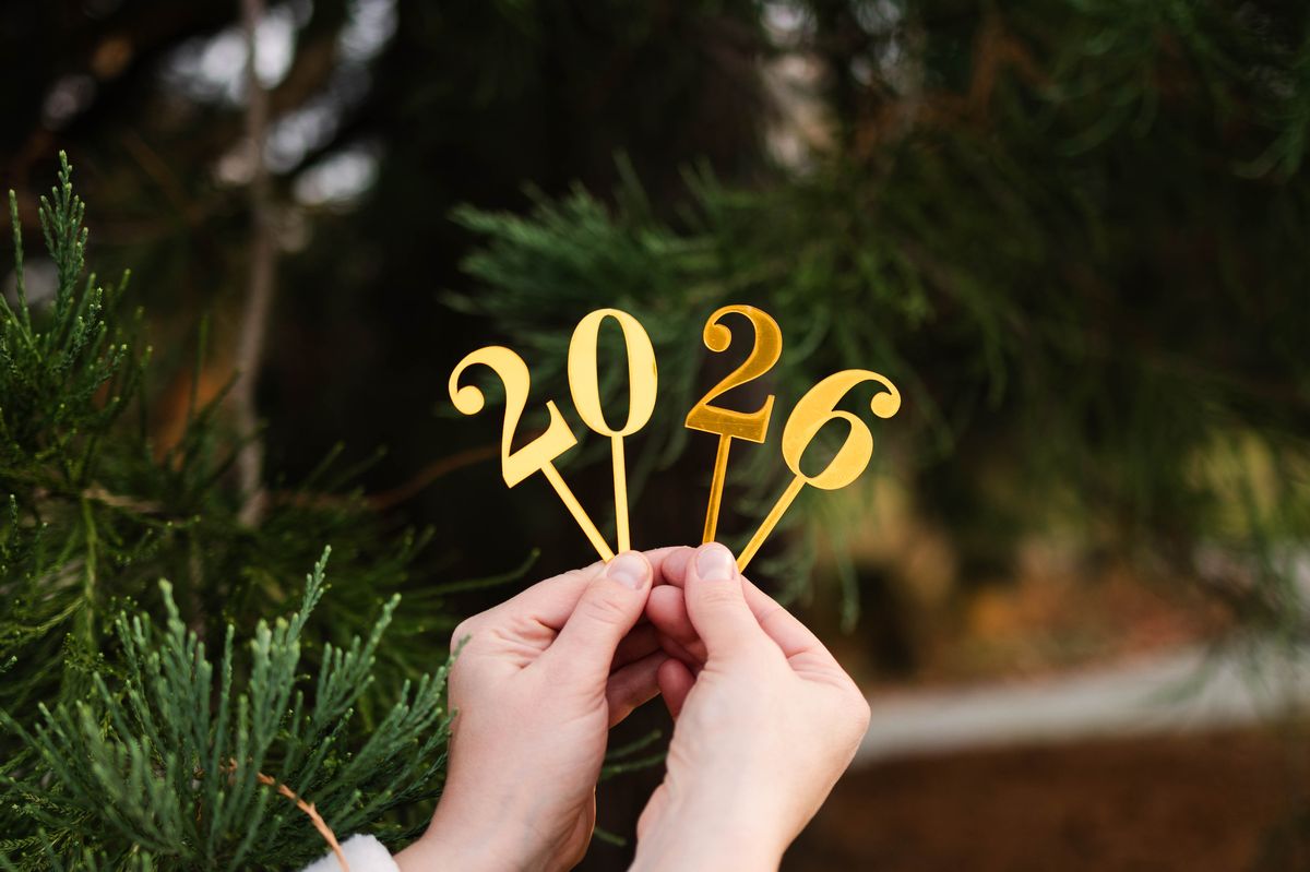 Close-up of a person holding golden “2026” numbers in front of evergreen branches outdoors. Festive New Year concept with warm natural light and holiday atmosphere.