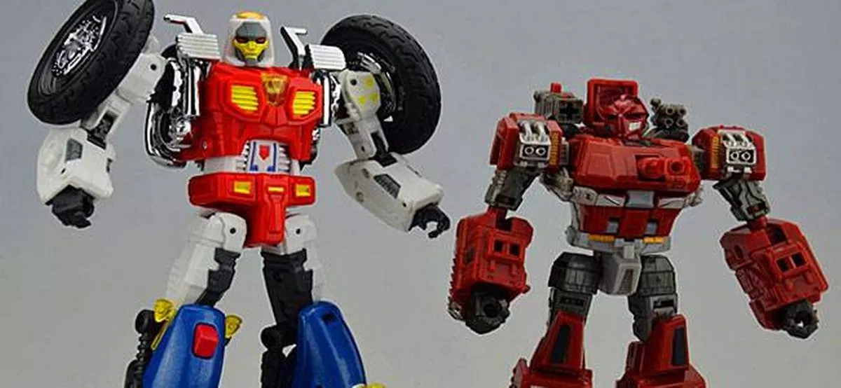 Two articulated toy robots are positioned next to each other on a gray background. The robot on the left is predominantly red and white with a black wheel on its right arm. The robot on the right is entirely red with intricate detailing and a prominent red chest plate.
