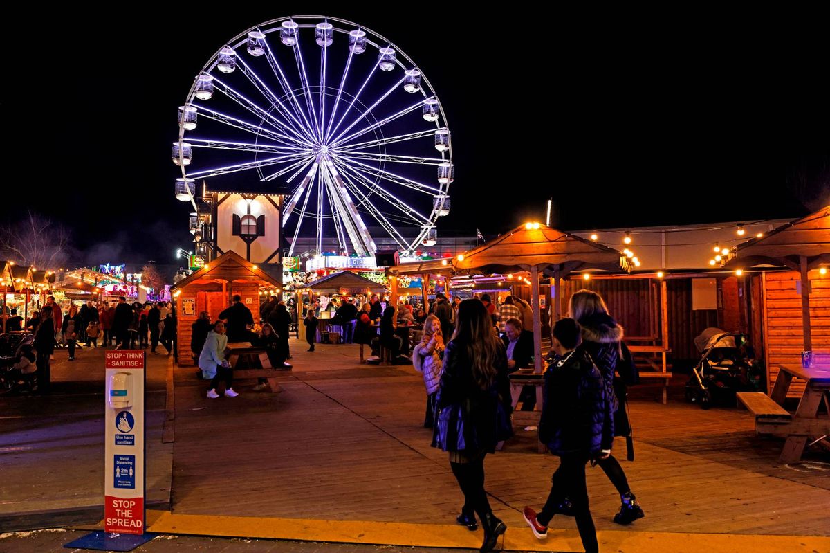 An illuminated night scene at a fairground, featuring a large Ferris wheel in the background. The fairground is bustling with activity, as people walk among various stalls and attractions. String lights and festive decorations adorn the area, creating a vibrant atmosphere.