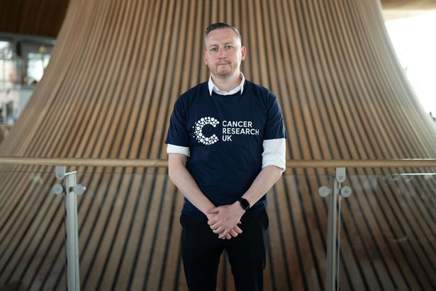 Jody, with fair hair and white skin, is pictured here in a Cancer Research t-shirt while posing in the Senedd in Cardiff Bay