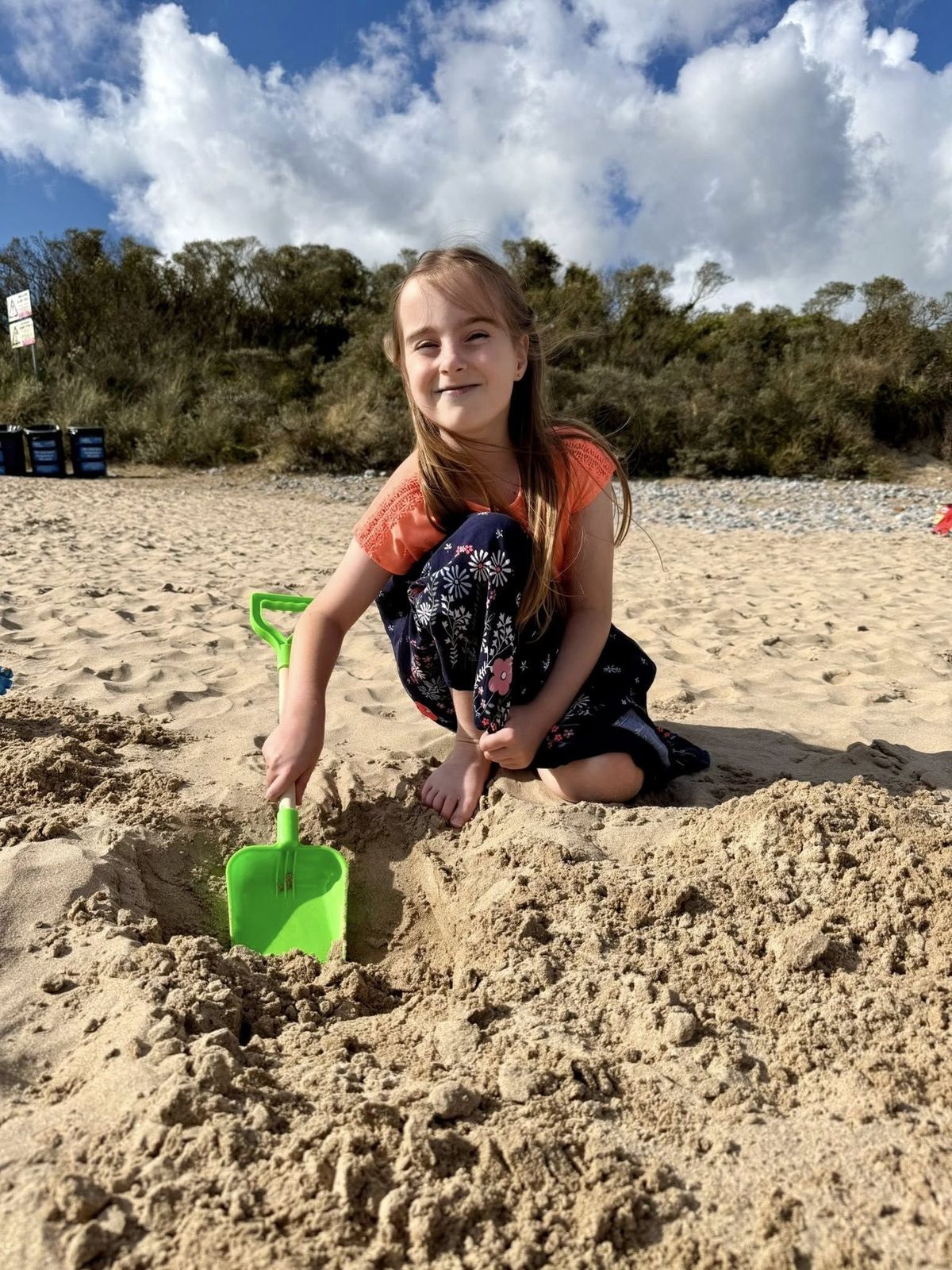 A little girl smiling as she digs into sand with a green spade