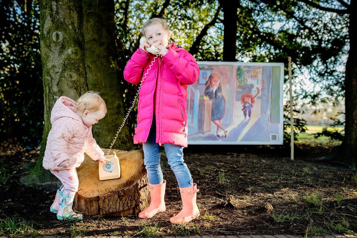Two young children in pink coats play with an old-fashioned telephone in front of an illustration in some woodland. The illustration is a cartoon-style pencil drawing of a woman using a phone while a child with a hockey stick stands in an open door behind her.
