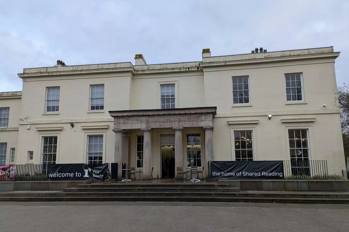 A cream-fronted Georgian mansion under a cloudy sky. Four Greek columns flank the door. The mansion has 11 large windows visible on the front and another wing visible to the left of the image. Banners outside read: "Welcome to The Reader. The home of Shared Reading."