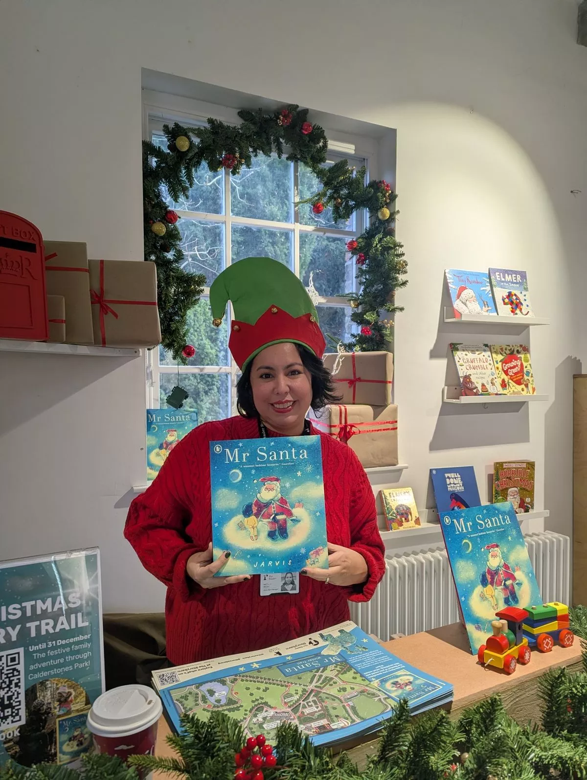 A woman with black hair wearing a red jumper and a green elf hat smiles while holding a book called "Mr. Santa". Behind her is a Christmas wreath and in front of her is a map of Calderstones Park.