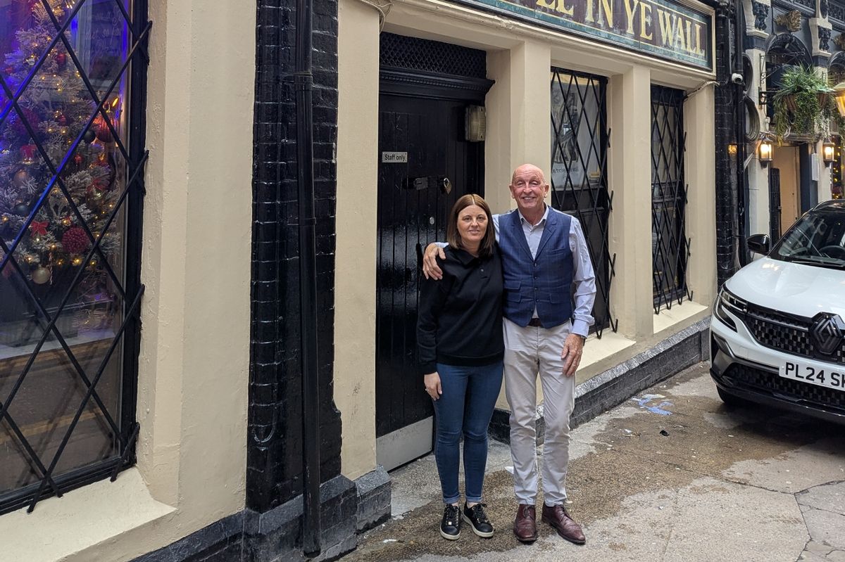 A man and woman smile standing in front of a pub with the sign: "Ye Hole in Ye Wall". The building is old, with visible wooden beams on the outside. A Christmas tree is lit up in the window.