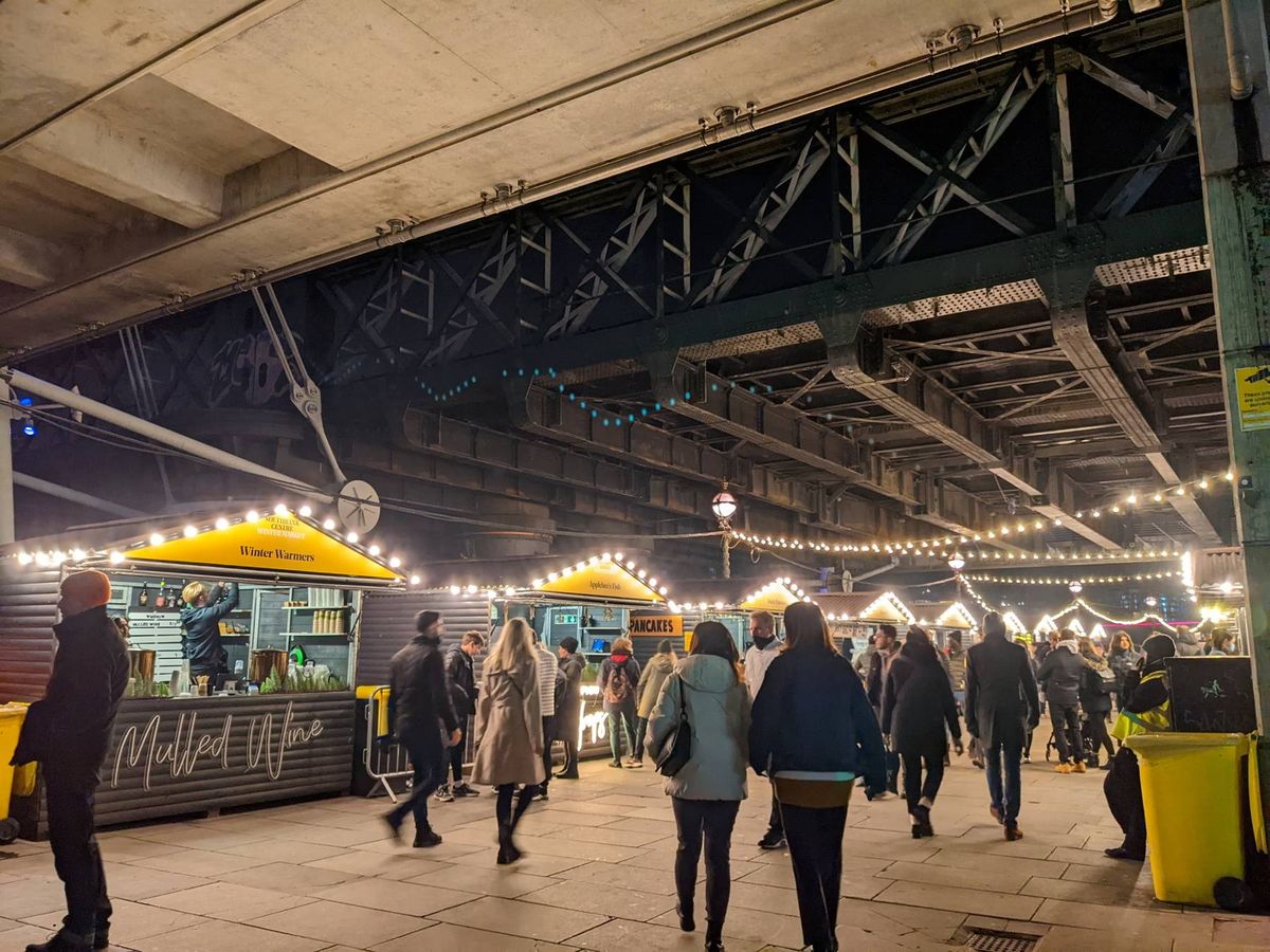 People browsing the busy Christmas Market under Hungerford Bridge and Golden Jubilee Bridges in Southbank at night London's