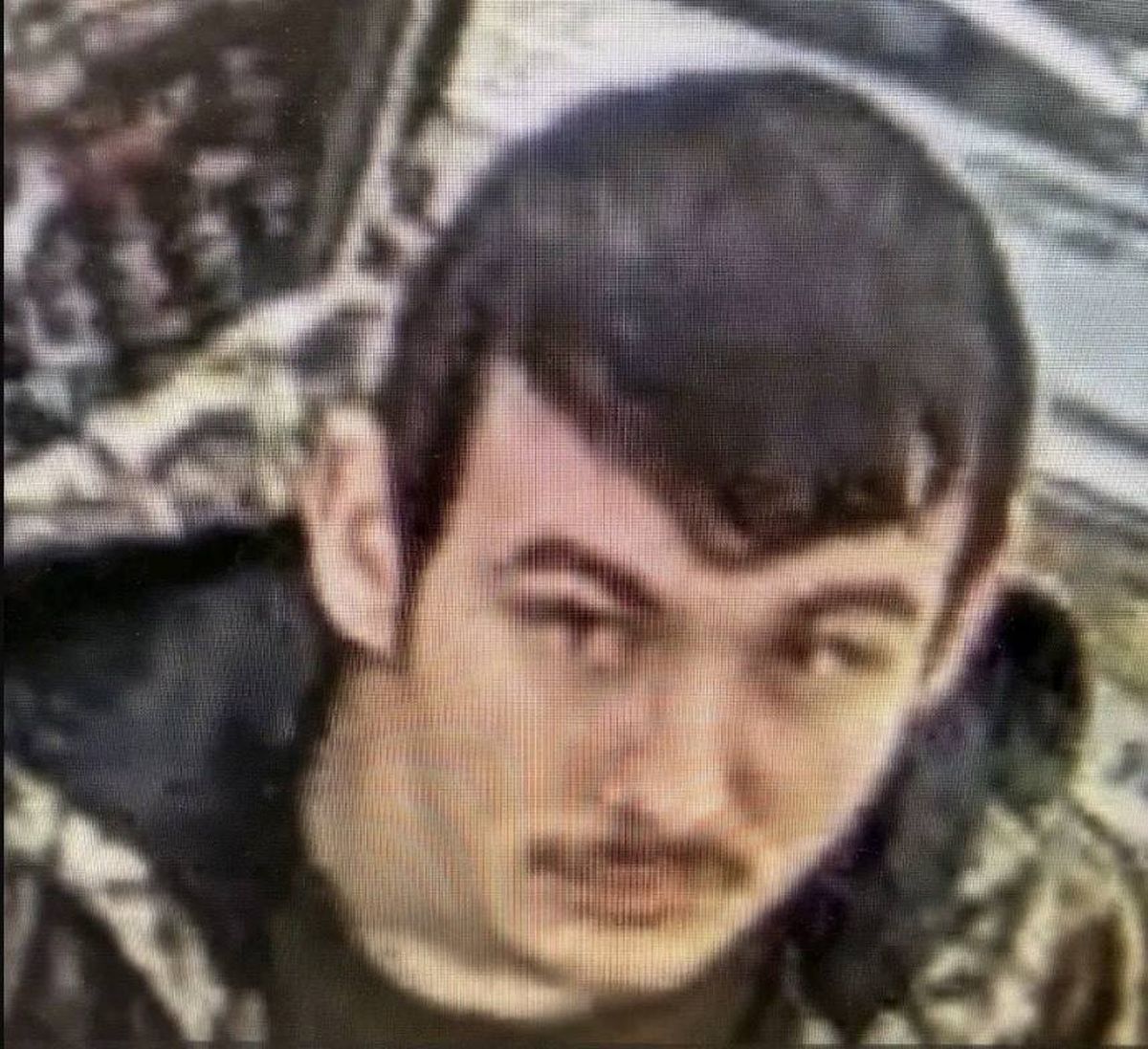 An image depicts a close-up portrait of an individual with short black hair and a mustache, wearing a black jacket against a camouflage background.