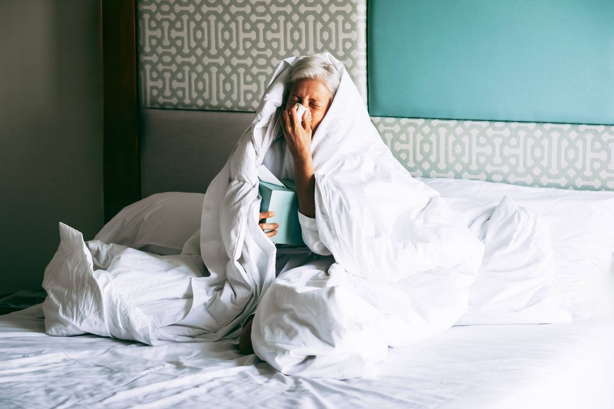 Woman with short gray hair sitting in bed under blanket, blowing nose into tissue. Holding mug and book. Cold or flu recovery concept.