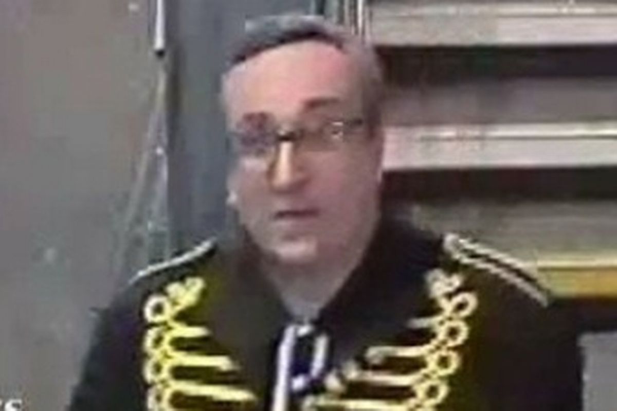 A white man in his late 30s or early 40s and of heavy build wearing glasses and a distinctive dark jacket with a gold design on it.