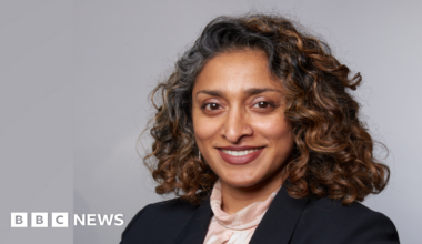 A head and shoulders image of Monisha Gower. She is wearing a black blazer over a light top. She is standing in front of a grey backdrop and smiling while looking into the camera.