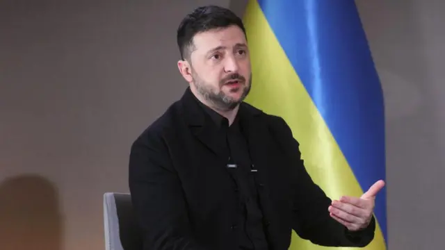 Zelenksy sat on chair in front of a Ukrainian flag