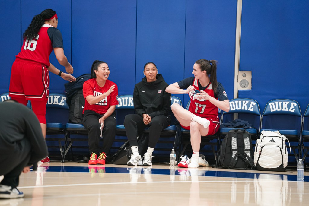 Indiana Fever guard Caitlin Clark sits on the bench before a Team USA practice. She talks with Natalie Nakase and JuJu Watkins as she sits with one leg crossed over her lap and holds one shoe in both hands.