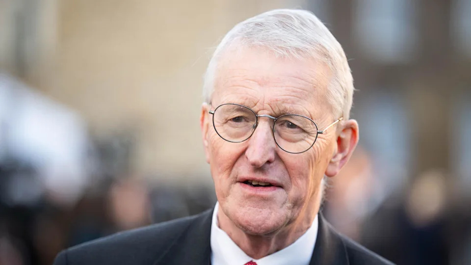 Secretary of State Hilary Benn is wearing round glasses with thin rims, and his thinning grey hair is swept to the side.