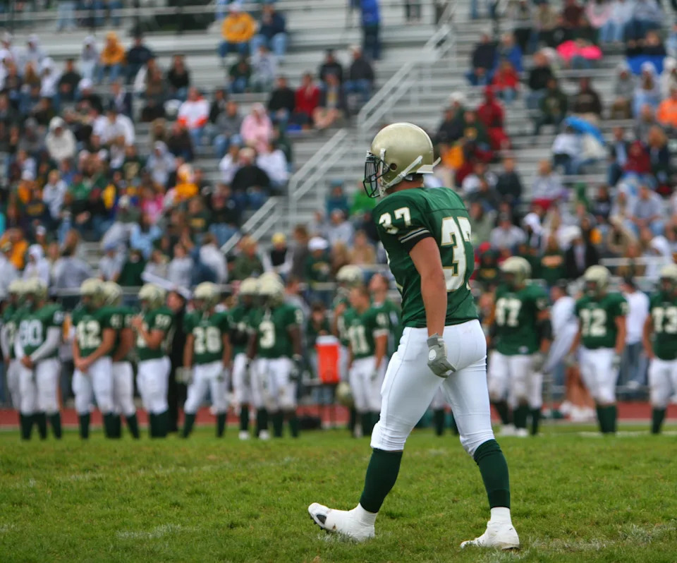 Football player in uniform walks on a field during a game, with a team and spectators in the background