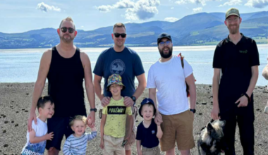Five men stood in a row, with four young boys and a dog stood in front of them, and one man holding a young girl in his arms. They are all looking at the camera and smiling. Behind them is a body of water, with mountains in the distance.