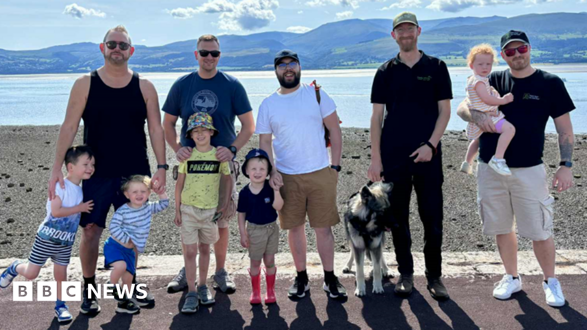 Five men stood in a row, with four young boys and a dog stood in front of them, and one man holding a young girl in his arms. They are all looking at the camera and smiling. Behind them is a body of water, with mountains in the distance.