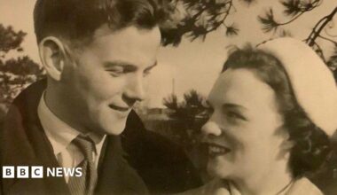 A black and white photo of a young man and woman looking at each other affectionately. The man is wearing an overcoat, a light shirt and a striped tie. The woman has short, curly dark hair under a light hat and is wearing lipstick.