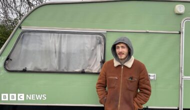 Jack Knight is standing in front of his green caravan, smiling at the camera. He is wearing a brown jacket.