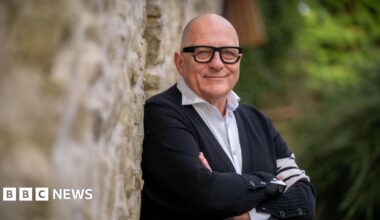 Hugh Wallace standing against a stone wall with his arms folded. He is bald, wearing glasses, a black cardigan with white shirt underneath.