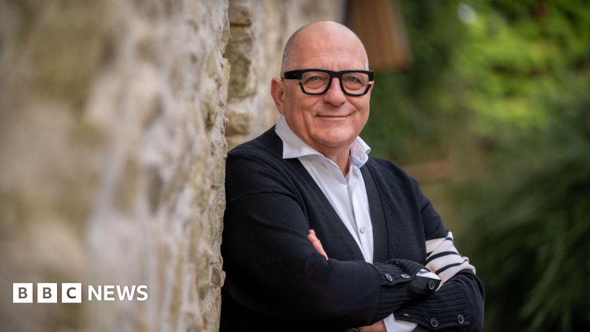 Hugh Wallace standing against a stone wall with his arms folded. He is bald, wearing glasses, a black cardigan with white shirt underneath.