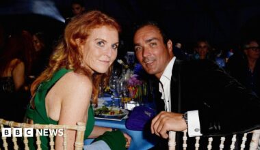 Sarah Ferguson and Manuel Fernandez sit at a table laden with glasses, plates and a blue table cloth. She wears a green dress, he wears a black velvet jacket and a white shirt open at the neck. They sit at wickerwork-style chairs, and are looking over their shoulders at the camera