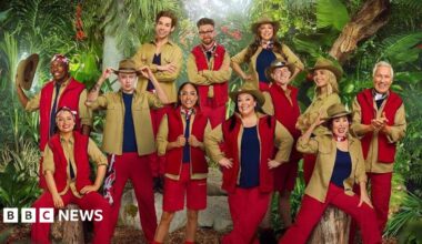 Twelve people can be seen wearing various outfits of red and khaki green, with some wearing large khaki hats. They are posed in front of a jungle backdrop as part of the announcement for the new cast of I'm a Celebrity...Get Me Out of Here!